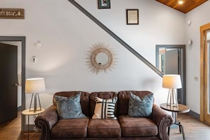 Ein modernes Wohnzimmer mit einem braunen Ledersofa, zwei Beistelltischen mit Lampen, einem dekorativen Spiegel im Sonnenstrahlendesign an der Wand und einer Treppe, die in einen anderen Raum führt.