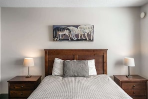 A bedroom with a wooden headboard, two bedside tables with lamps, a patterned bedspread, and a painting of wolves on the wall.