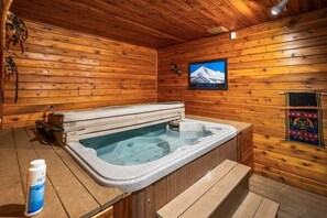 A wooden cabin interior with a hot tub, wooden walls, a mounted television, and a window with a mountain view.