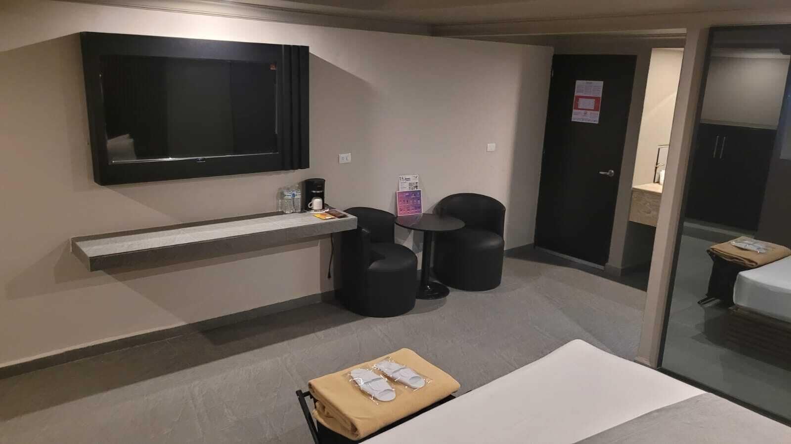 A modern hotel room with a flat-screen TV mounted on the wall, a small table with a cup and a bottle, two black ottomans, a bed with a white mattress, and a glass partition separating the room from another space.