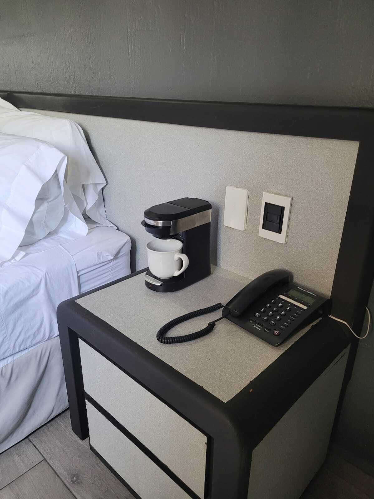 A compact hotel room with a single bed, a small desk with a coffee maker, a telephone, and a power outlet.