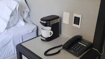 A compact hotel room with a single bed, a small desk with a coffee maker, a telephone, and a power outlet.