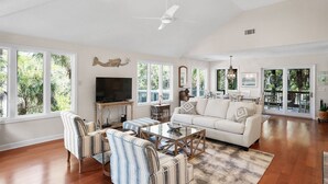 A spacious living room with a large sofa, striped armchairs, a coffee table, and a television. There are large windows providing natural light and views of greenery outside. - Pets  Considered, Community Pool, Lagoon View, Golf Cart Included 875 Salt Cedar Lane (Fripp Island)