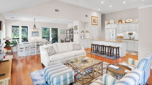 A spacious living area with a white sofa, striped ottoman, and glass coffee table. A kitchen with white cabinets and a dining area with a wooden table and chairs are visible. - Pets  Considered, Community Pool, Lagoon View, Golf Cart Included 875 Salt Cedar Lane (Fripp Island)