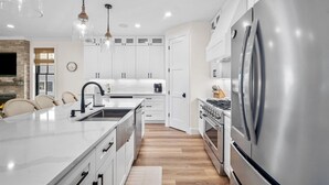 A modern kitchen with white cabinetry, stainless steel appliances, and a large island with a sink. - Shuck Yeah - Brand New, Panoramic Marsh and Golf Views, Golf cart included 92 Davis Love (Fripp Island)