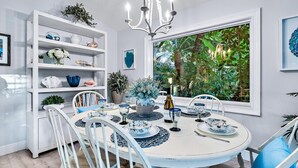 A dining area with a round table, white chairs, and a chandelier. There is a window with a view of greenery outside.