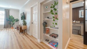 A modern living room with a wooden floor, a white door, a bookshelf with plants, and a flat-screen TV on the wall. - Cozy Oasis Apt - In Med Center & NRG (Houston)