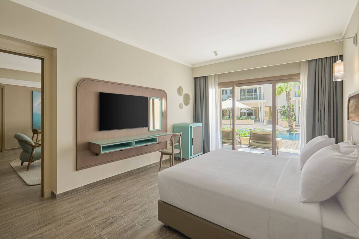 A hotel room with a large bed, a flat-screen TV mounted on the wall, a desk with a chair, and a view of the pool area.