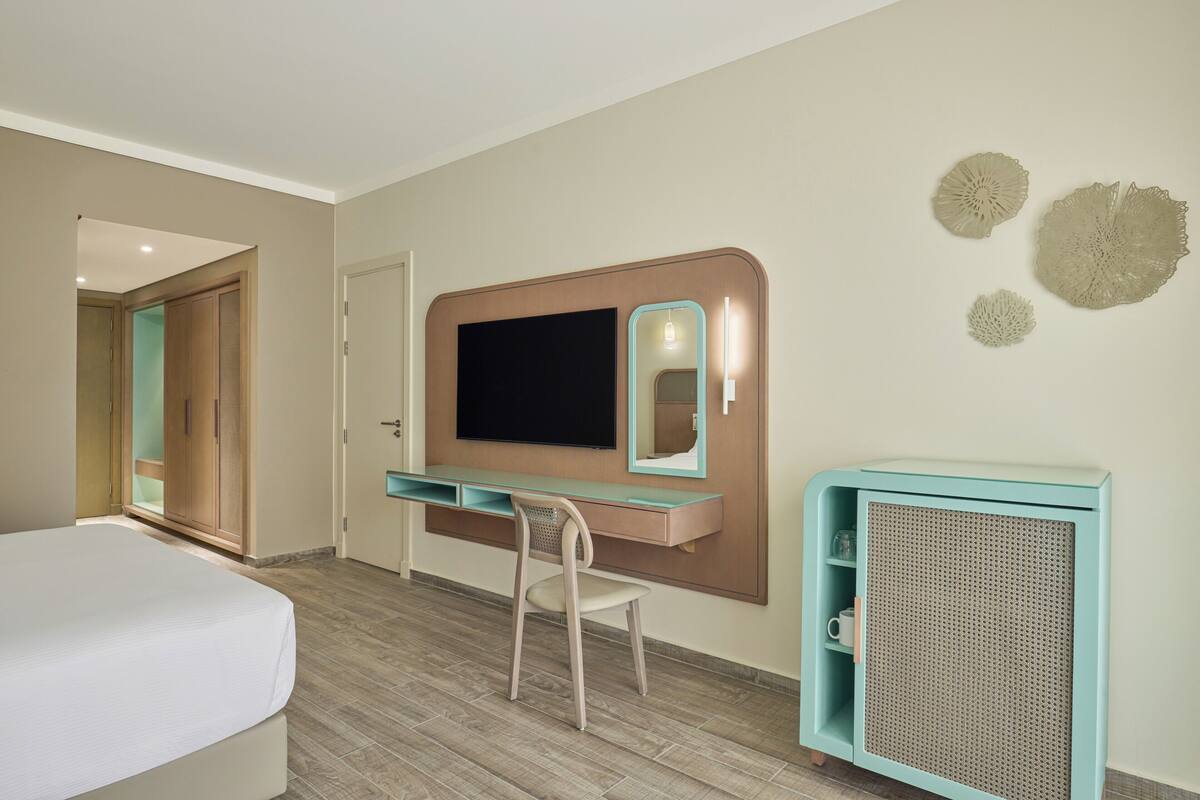 A hotel room with a bed, a desk with a flat-screen TV, a chair, a small cabinet, and decorative wall art.