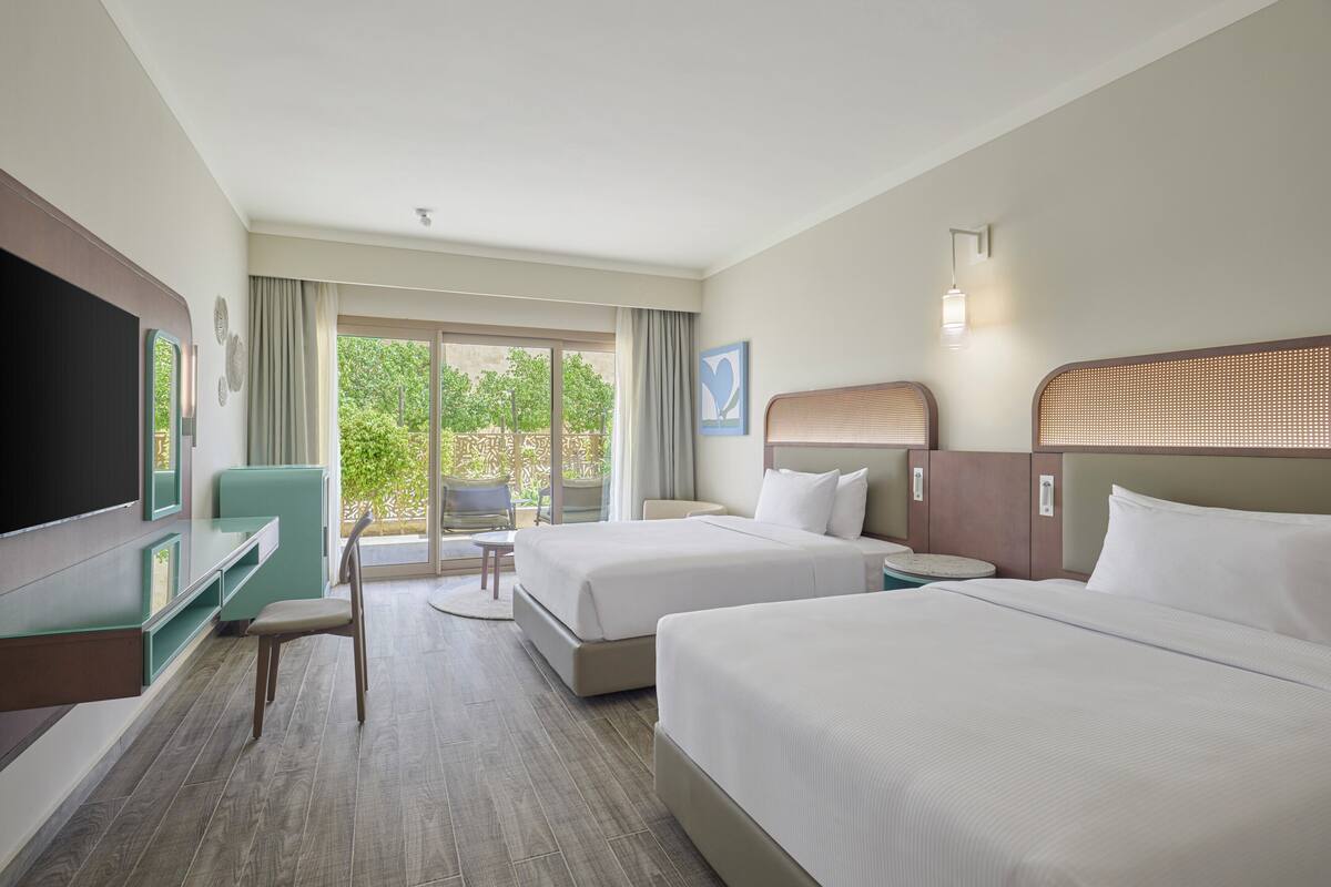 A hotel room with two beds, a desk, a TV, and a view of the outdoors.