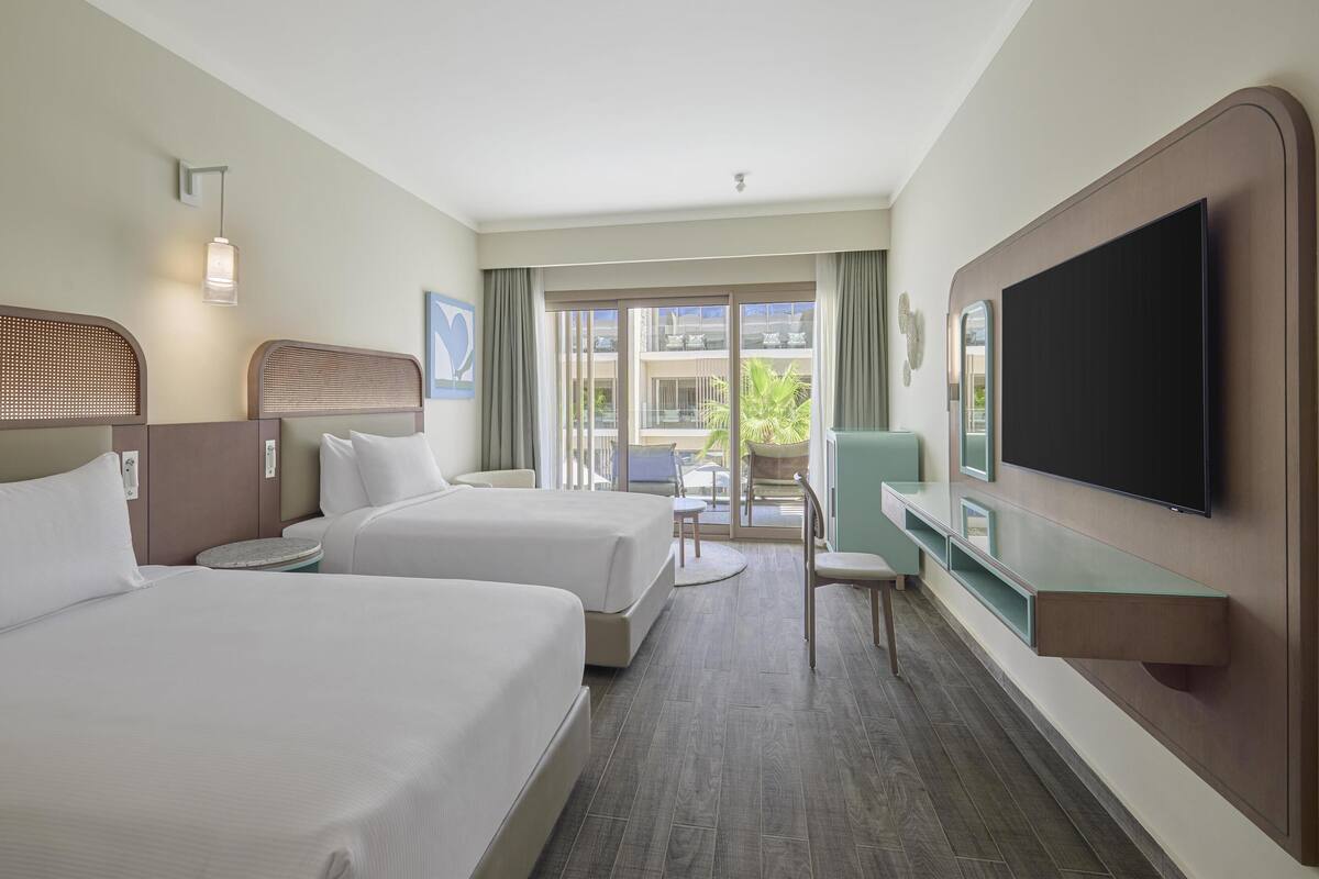 A hotel room with two beds, a TV, a desk, and a view of the outside.