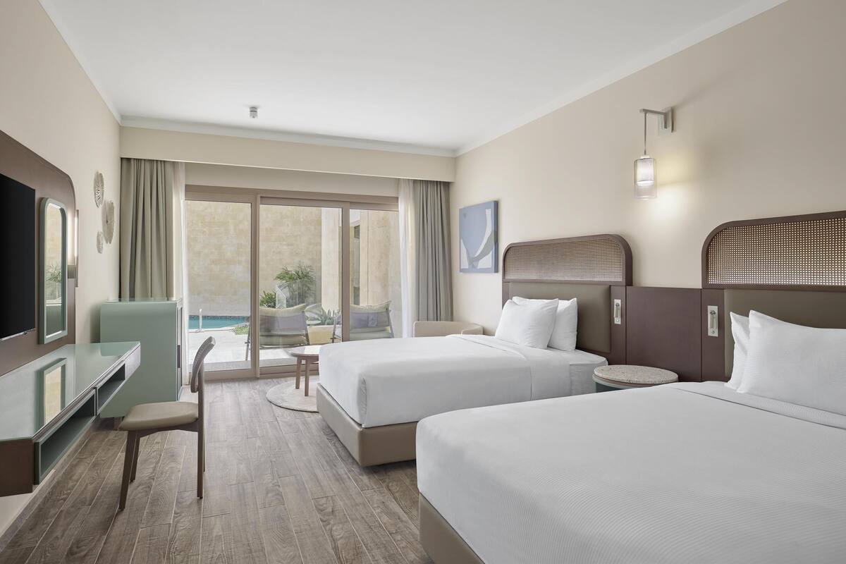 A hotel room with two beds, a desk, a chair, and a TV. There is a balcony with a view of the outdoors.