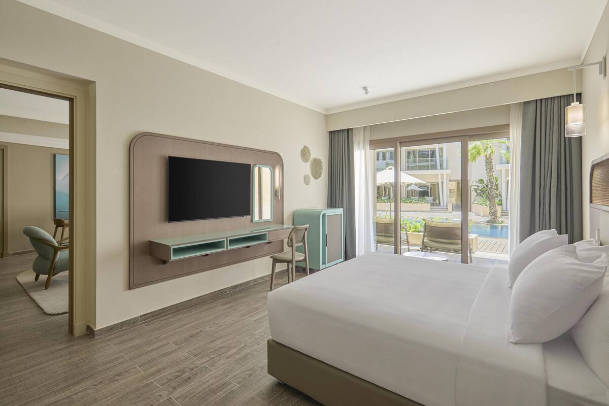 A hotel room with a large bed, a TV mounted on the wall, a desk, and a view of the outdoor pool area.