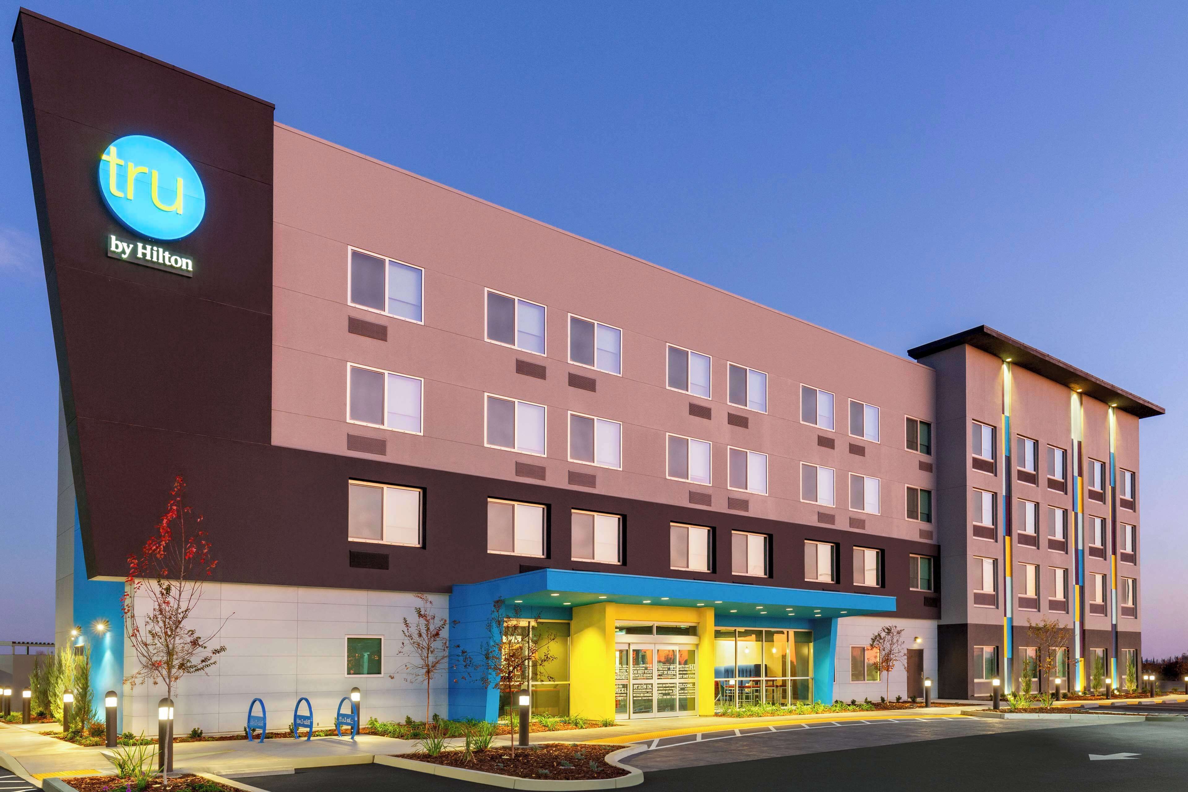 A modern hotel building with a prominent 'tru by Hilton' sign, multiple windows, and a well-lit entrance area.