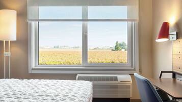 A hotel room with a large window, a bed with a patterned bedspread, a desk with a chair, and a wall-mounted lamp.