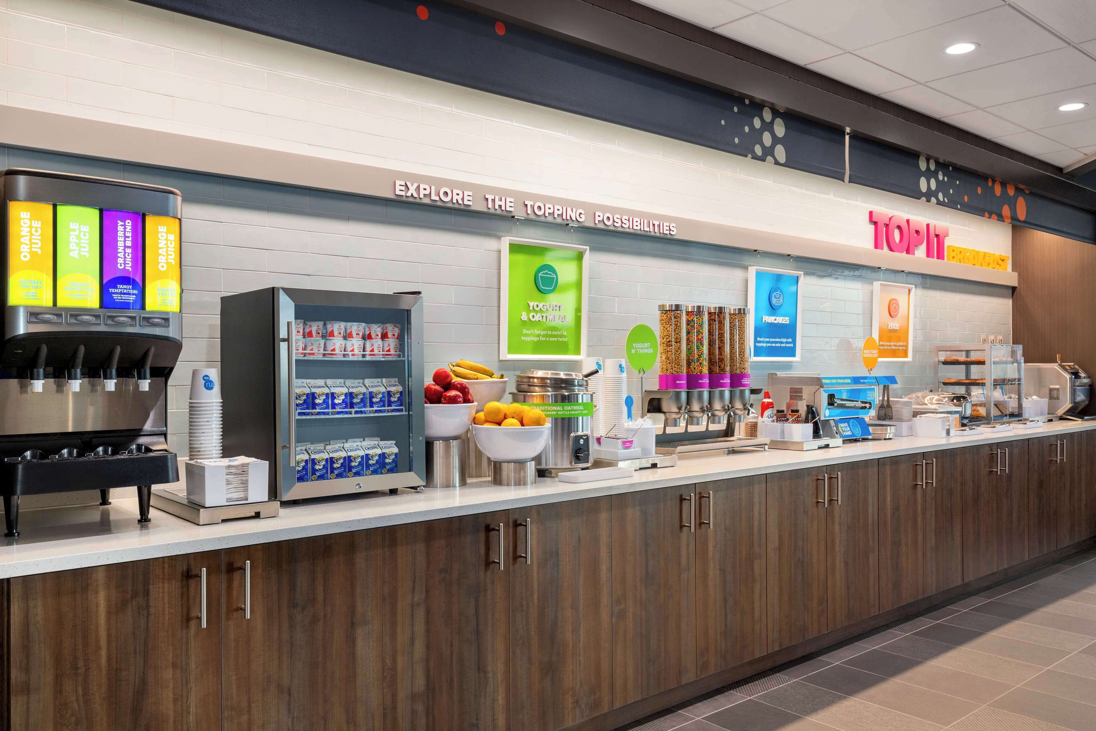 Un moderno espacio para comidas con dispensador de zumo, estación de yogur y avena, y una variedad de alimentos.