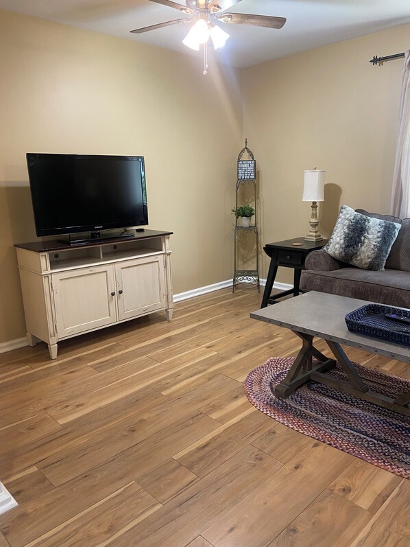 A living room with a flat-screen TV on a cabinet, a ceiling fan, a side table with a lamp, a sofa with a patterned throw pillow, and a coffee table.