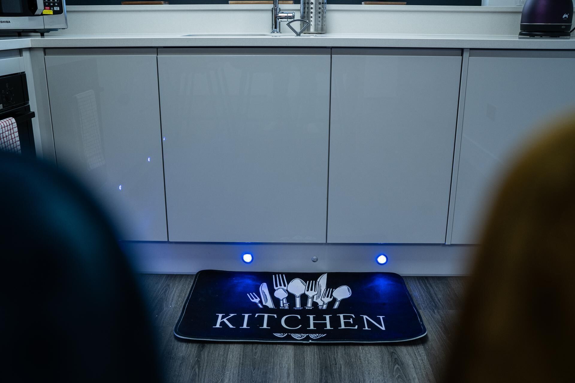 A kitchen area with a stainless steel sink, a microwave, and a black kitchen mat with utensils and the word "KITCHEN" on it.