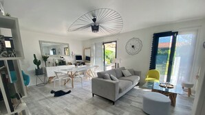 Una sala moderna con un sofá gris, un sillón amarillo y una mesa de centro de vidrio. Se observa un comedor con una mesa y sillas blancas. También se distingue un espejo grande y un adorno geométrico en la pared.