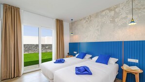 A hotel room with two beds, a large window with curtains, a blue headboard, a small bedside table, and a wall with a floral pattern. - Villa Tirma by Villa Plus (Playa Blanca)