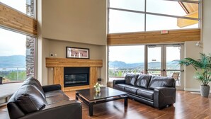 A spacious living room with a fireplace, leather sofas, and large windows offering a view of the mountains. - Top Floor Lake View Condo at Copper Sky (West Kelowna)