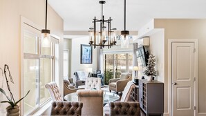 A spacious living room with a large chandelier, comfortable seating, and a view of the outdoors. - Top Floor Lake View Condo at Copper Sky (West Kelowna)