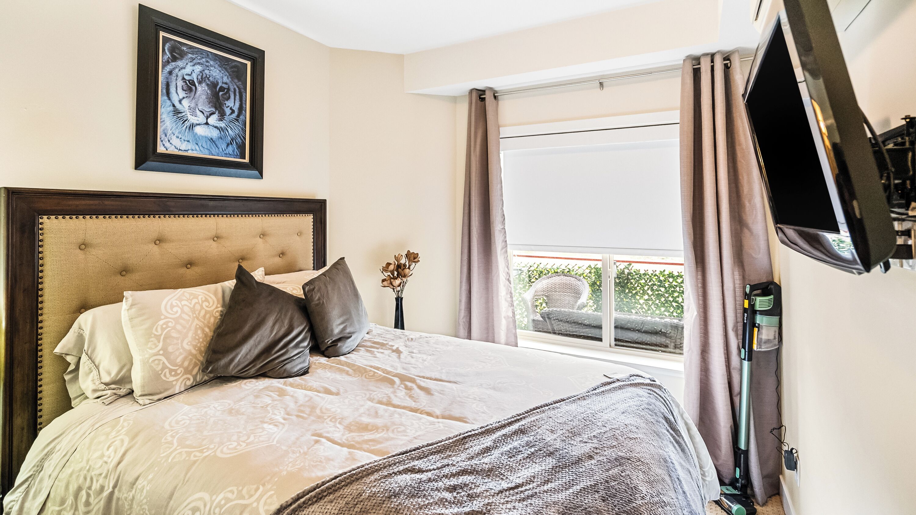 A bedroom with a large bed, a framed picture on the wall, a window with curtains, and a television mounted on the wall.