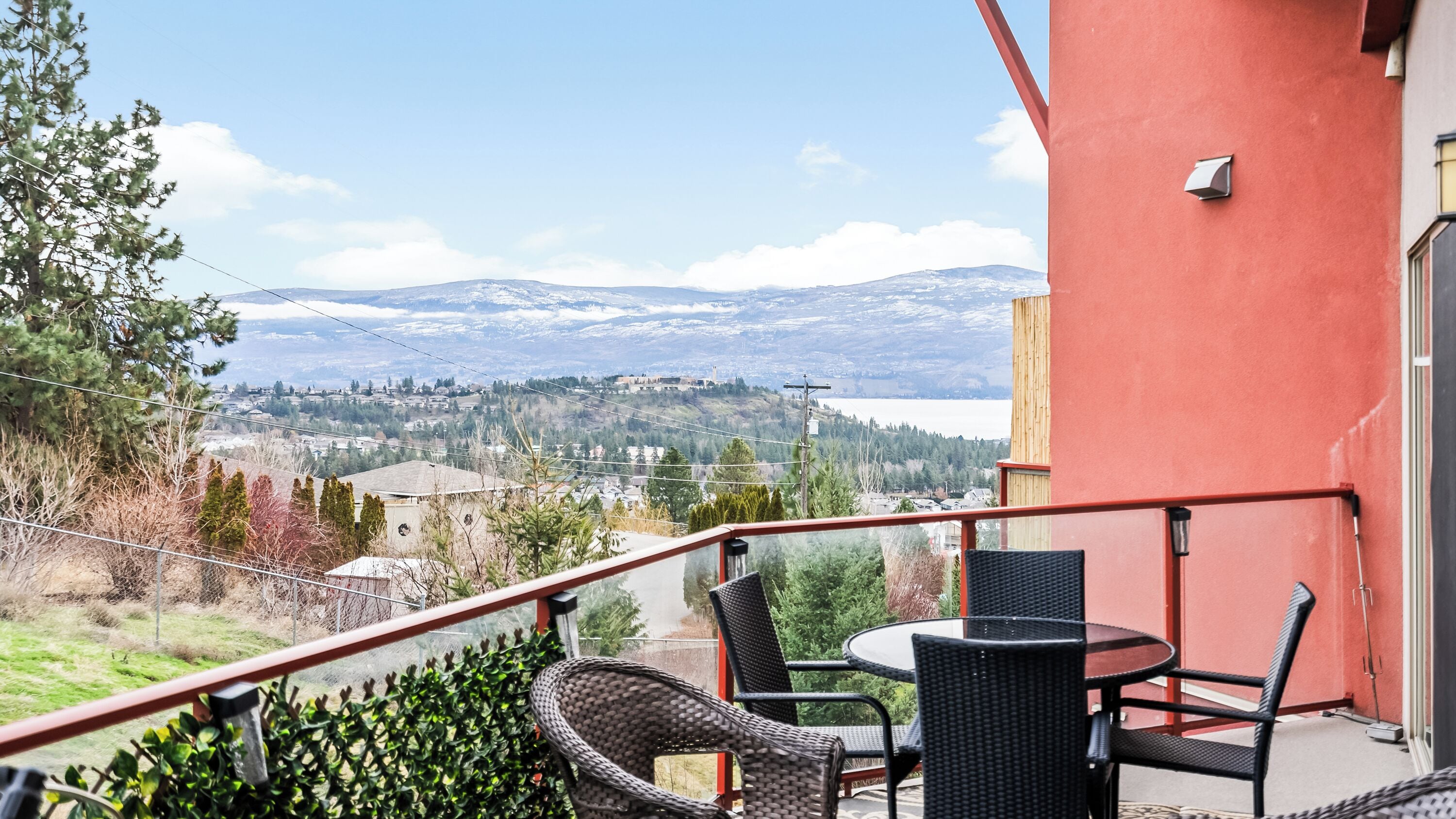 Un balcon doté de fauteuils et d’une table en osier, offrant une vue sur un paysage montagneux avec un lac et des bâtiments.