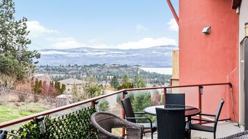 Un balcon doté de fauteuils et d’une table en osier, offrant une vue sur un paysage montagneux avec un lac et des bâtiments.
