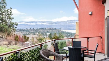 Un balcón con sillas de mimbre y una mesa, con vistas a un paisaje montañoso con un lago y edificios.