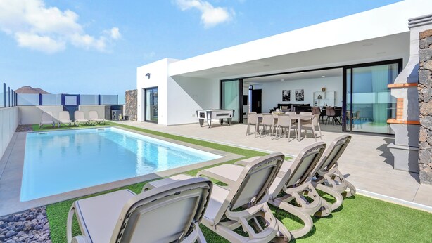 Un espace piscine extérieur moderne, doté de chaises longues, d’un coin repas avec tables et chaises, et d’un mur en pierre.