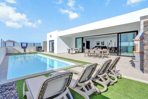 Un área de piscina moderna al aire libre con sillas de descanso, un comedor con mesas y sillas, y un muro de piedra.