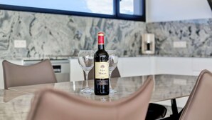 Une bouteille de vin est posée sur une table, accompagnée de deux verres, dans une cuisine moderne dotée d’un plan de travail en marbre et de grandes fenêtres.