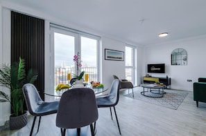 A modern living room with a dining area, large windows, a flat-screen TV, and a city view. - Cosy 2 bed Apt n/ City Centre & Mediacity (Salford)