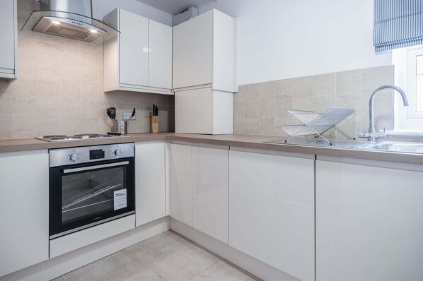 A modern kitchen with white cabinetry, a stainless steel range hood, a built-in oven, and a sink with a metal drying rack.