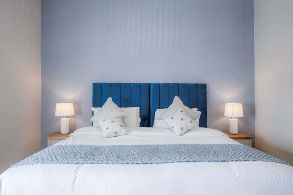 Una cama bien hecha con ropa blanca y detalles azules, flanqueada por dos lámparas de noche sobre mesitas de madera.
