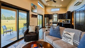 A spacious living room with a large window, a fireplace, a sofa with decorative pillows, a coffee table with books, and a view of a grassy field.