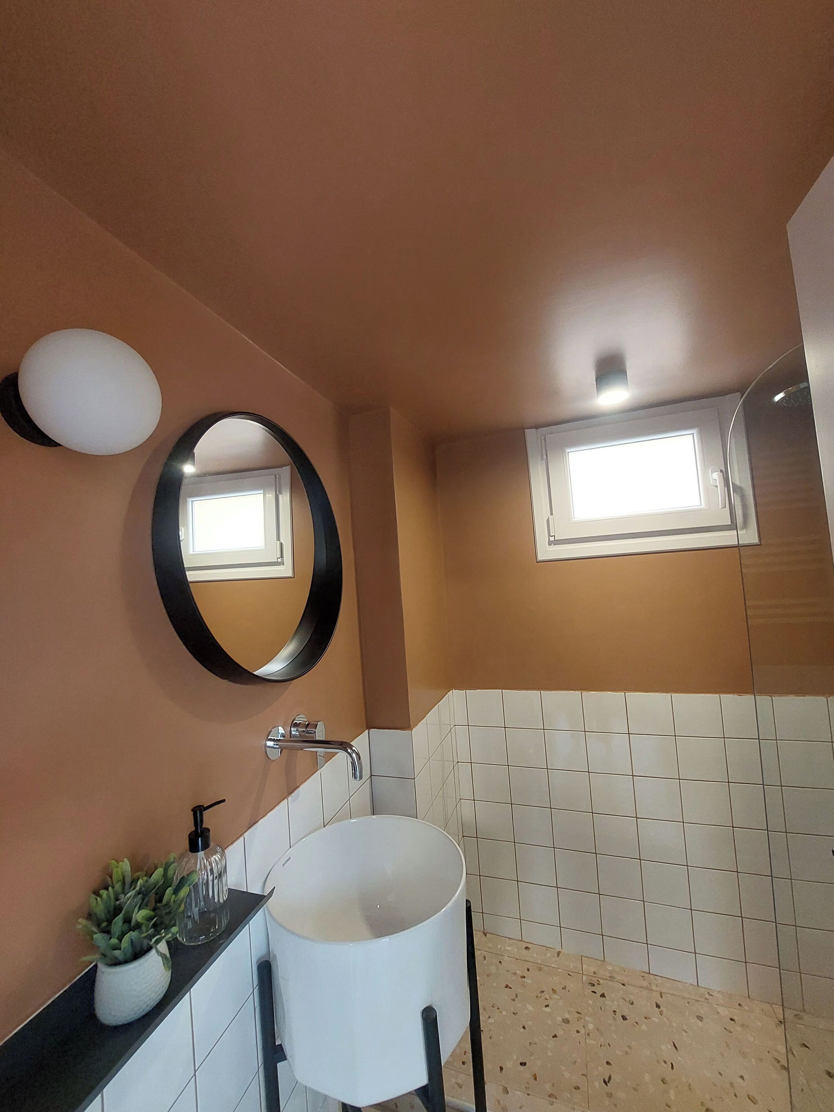 A bathroom with a white sink, a round mirror, a potted plant, and a small window.