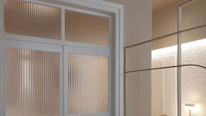A room with a sofa, a round ottoman, a white wardrobe with frosted glass panels, and a mirror reflecting a lit lamp and chair. - Terrazzo City Apts (Heraklion)