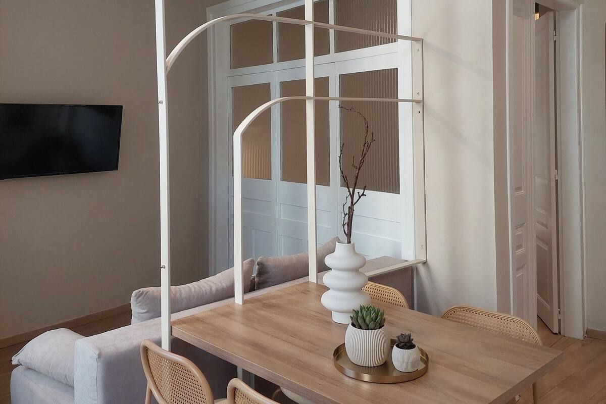 Een moderne eetruimte met een houten tafel, vier stoelen, een witte vaas met een tak en een aan de muur bevestigde televisie.