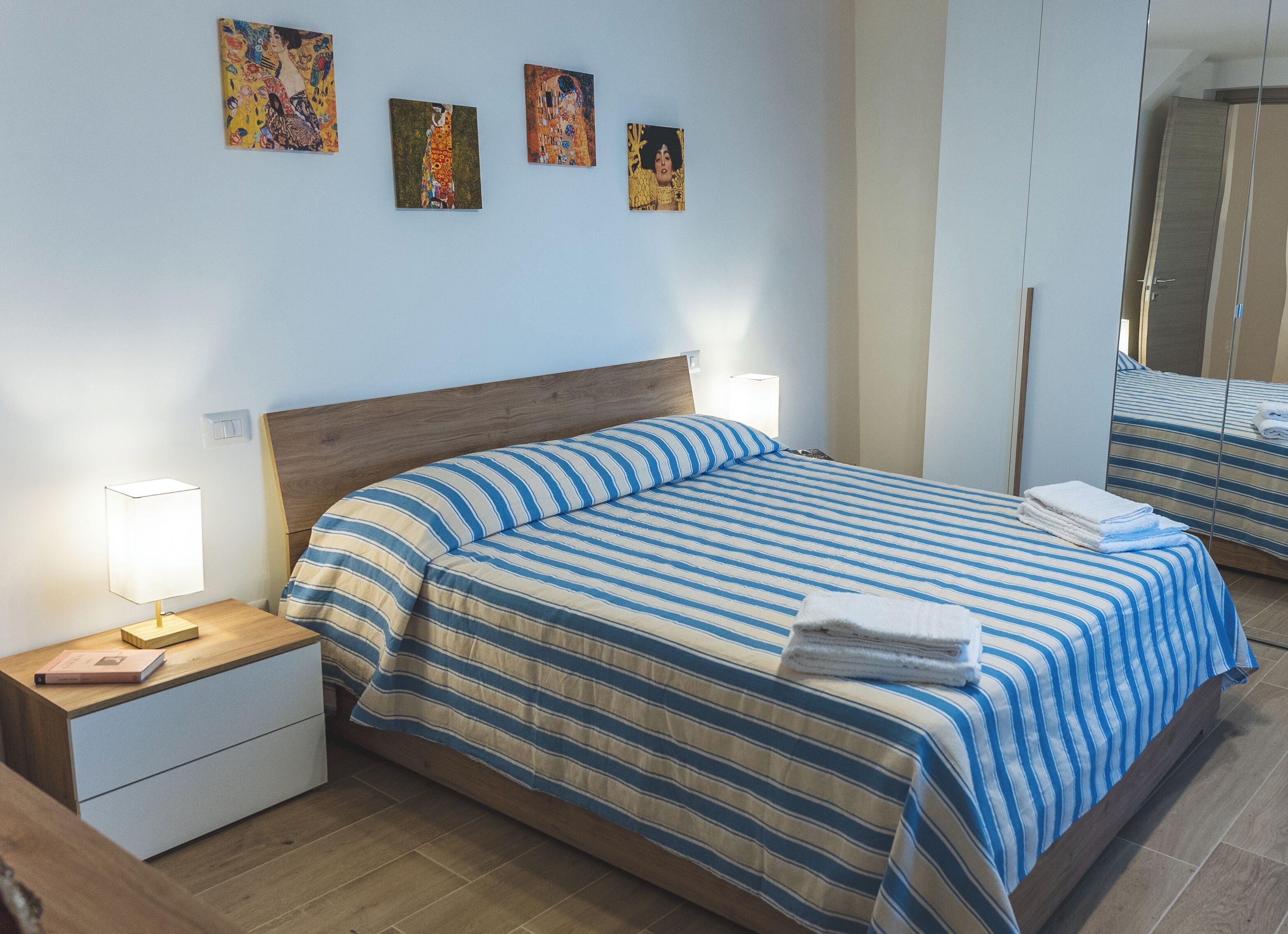 A neatly made bed with striped bedding, a wooden headboard, bedside tables with lamps, and a mirror reflecting the bed.