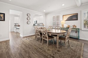A dining area with a round table, chairs, a rug, a kitchen in the background, and a painting on the wall.