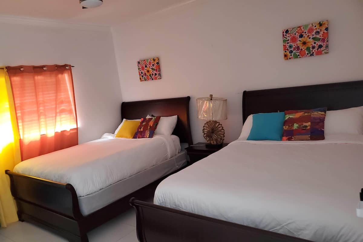 Two single beds with wooden frames, white bedding, and colorful pillows. There is a nightstand with a lamp and a small decorative item between the beds.