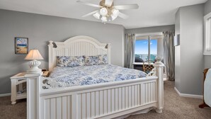 A bedroom with a bed, a nightstand, a lamp, a ceiling fan, and a window with curtains. - Indulge in stunning views from this expansive oceanfront condo featuring a large balcony and pool (North Myrtle Beach)