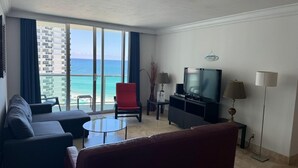 Un salón moderno con un televisor grande, asientos cómodos, una pequeña mesa de centro y vistas al océano a través de amplios ventanales.