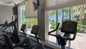 Un gimnasio con cintas de correr y elípticas, ventanas amplias con vistas a palmeras y un edificio.
