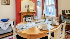 A dining area with a round wooden table set for four, surrounded by white chairs. A mirror hangs on the wall above the table, and a window with curtains is visible to the right.