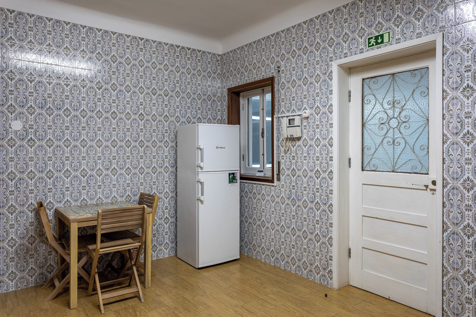 Uma cozinha compacta com paredes revestidas de azulejos estampados, uma geladeira branca, uma mesa de jantar de madeira com cadeiras e uma porta com painel de vidro decorativo.