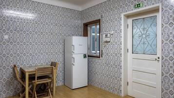 Uma cozinha compacta com paredes revestidas de azulejos estampados, uma geladeira branca, uma mesa de jantar de madeira com cadeiras e uma porta com painel de vidro decorativo.