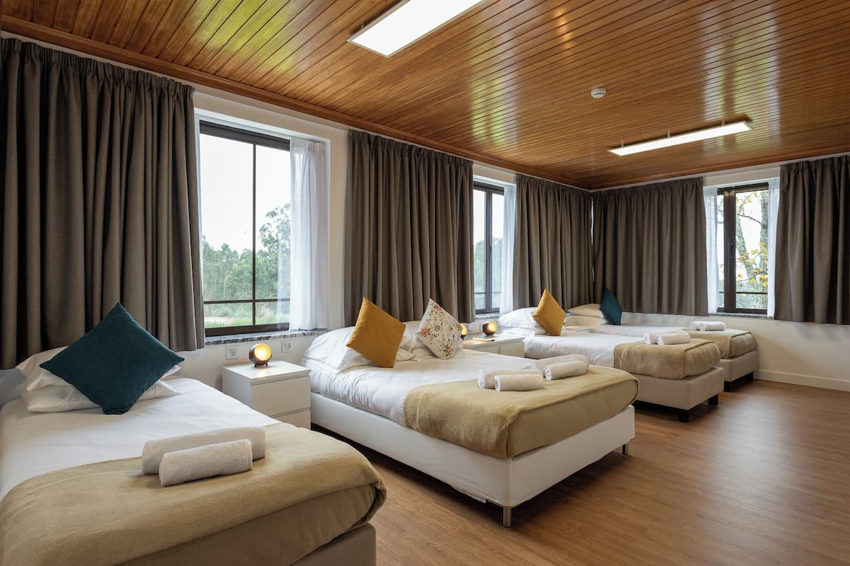 A hotel room with three beds, wooden ceiling, large windows, and a view of greenery.